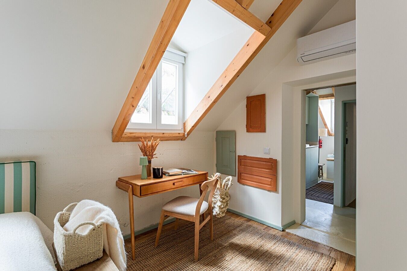 A room with a sloped ceiling, a desk with a chair, a small window, and a door leading to another room.