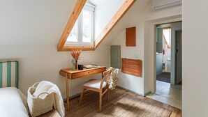Ein Zimmer mit schrägen Wänden, einem Schreibtisch mit Stuhl, einem kleinen Fenster und einer Tür zu einem weiteren Raum.