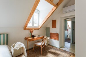 Ein Zimmer mit schrägen Wänden, einem Schreibtisch mit Stuhl, einem kleinen Fenster und einer Tür zu einem weiteren Raum.