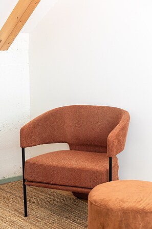Ein einzelner, braun bezogener Sessel mit gebogener Rückenlehne und passendem Hocker, platziert auf einem gemusterten Teppich vor einer weißen Wand mit Holzbalkendecke.