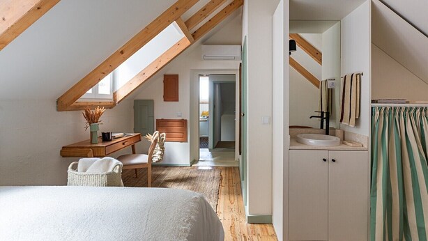 Ein Schlafzimmer mit schrägen Wänden, einem Bett, einem Schreibtisch, einem Stuhl, einer kleinen Kommode und einem Badezimmer mit Waschbecken und Spiegel.