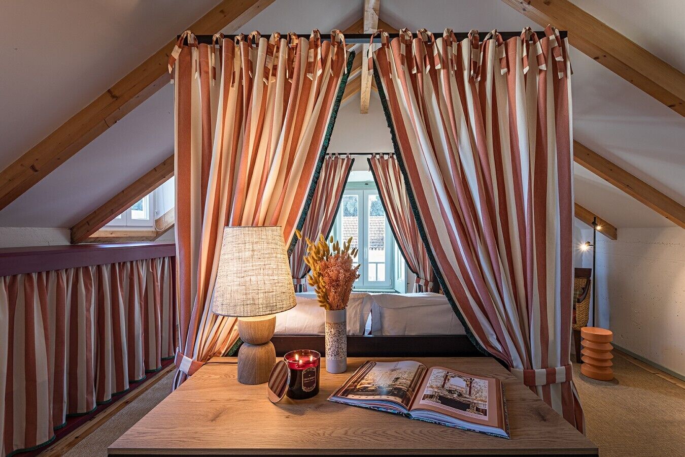 Ein gemütliches Schlafzimmer im Dachgeschoss mit schrägen Wänden, gestreifte Vorhänge, einer Nachttischlampe, einer Vase mit Trockenblumen und einem Buch auf dem Boden.