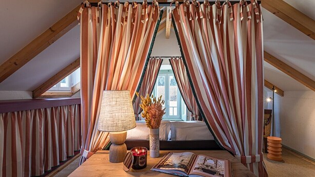 Een gezellig slaapkamer op zolder met een schuine kap, gestreepte gordijnen, een bedlampje, een vaas met gedroogde bloemen en een boek op de vloer.