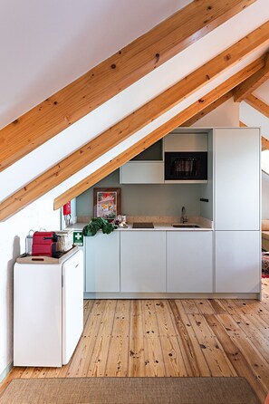 A compact kitchen with white cabinets, a microwave, and a sink under a sloped ceiling with exposed wooden beams. - The Old Net Studio in Porto Brandão (Caparica)