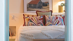 A bedroom with a bed, pillows, a framed picture, a bedside lamp, a wicker basket, and a wall-mounted air conditioner. - Tanoeiro's Cottage in Porto Brandão (Caparica)