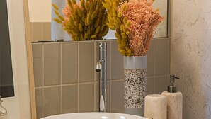 Uma casa de banho com uma pia branca, um espelho, um dispensador de toalhas e um vaso com flores secas.