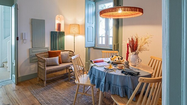 Uma área de refeições com mesa redonda, cadeiras, luminária e um vaso com flores secas.