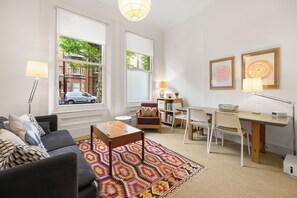 A living room with a sofa, coffee table, and patterned rug. There are two windows with a view of buildings and trees. A desk with chairs and a lamp is in the corner. - Chelsea Apartment | 2 Bedrooms | Spacious Garden (London)