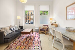 A living room with a sofa, coffee table, patterned rug, dining table, chairs, and a view of buildings through large windows. - Chelsea Apartment | 2 Bedrooms | Spacious Garden (London)