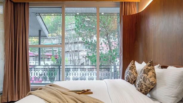 Una habitación de hotel con un ventanal grande, una cama con ropa de cama blanca, un plaid marrón y almohadas decorativas.