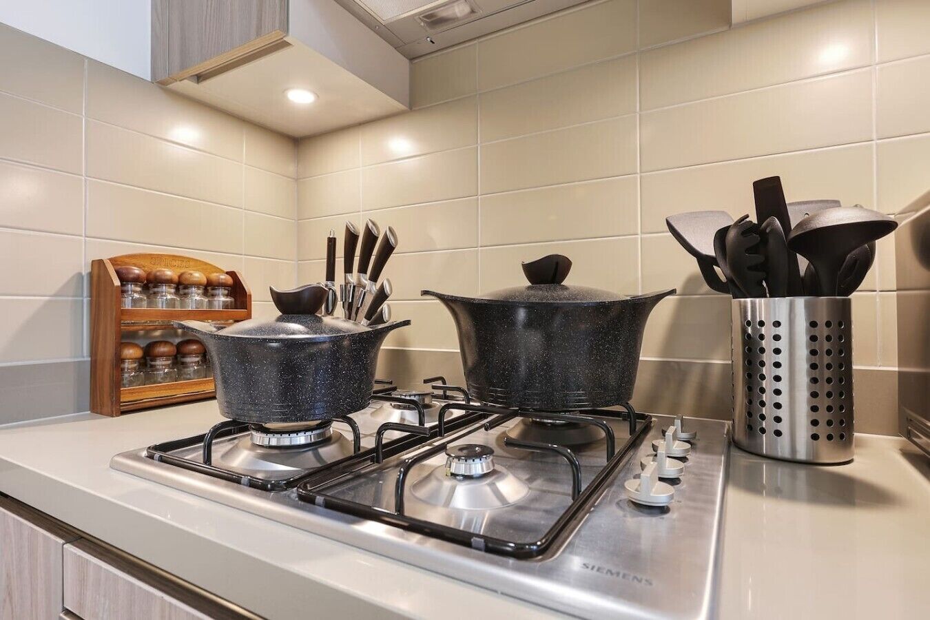 Une cuisine équipée de deux cuisinières à gaz, une casserole sur la cuisinière de gauche et une autre sur celle de droite. On y trouve un étagère à épices avec des pots et un porte-ustensiles contenant des cuillères et des spatules.