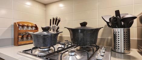 Une cuisine équipée de deux cuisinières à gaz, une casserole sur la cuisinière de gauche et une autre sur celle de droite. On y trouve un étagère à épices avec des pots et un porte-ustensiles contenant des cuillères et des spatules.