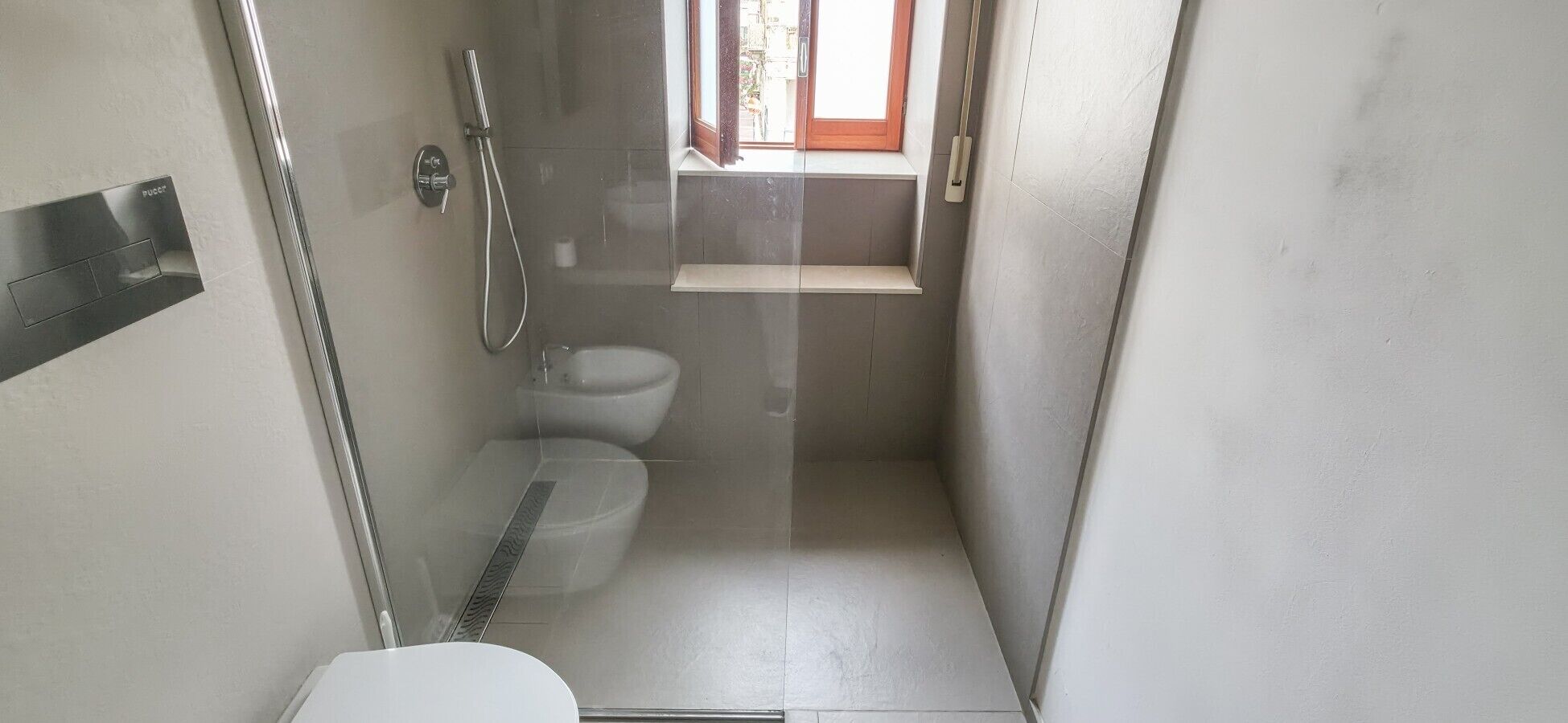 A small, narrow bathroom with a toilet, a shower, and a window.