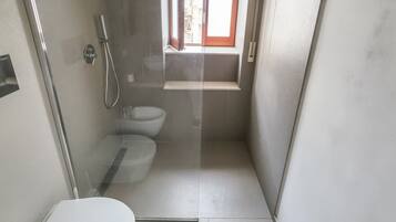 A small, narrow bathroom with a toilet, a shower, and a window.