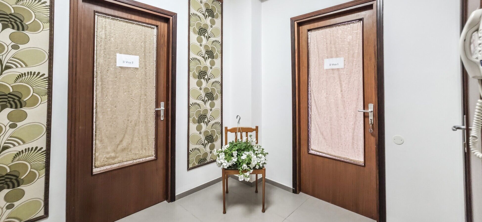 A hallway with two wooden doors labeled 'Il Vico 1' and 'Il Vico 2', a small wooden chair with a floral arrangement, and a patterned wallpaper on the left wall.