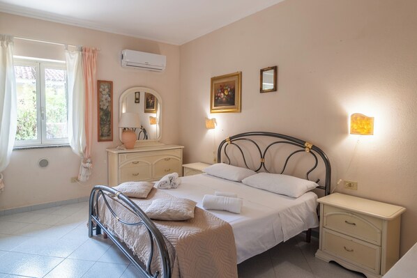 A bedroom with a bed, bedside tables, a dresser, a mirror, and a window with curtains. - San Nikola House (Olbia)