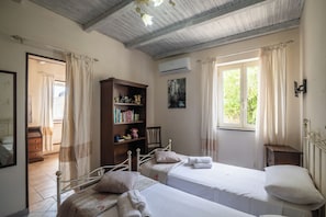 A bedroom with two beds, a bookshelf, a chair, a small table, an air conditioning unit, and a window with curtains. - San Nikola House (Olbia)