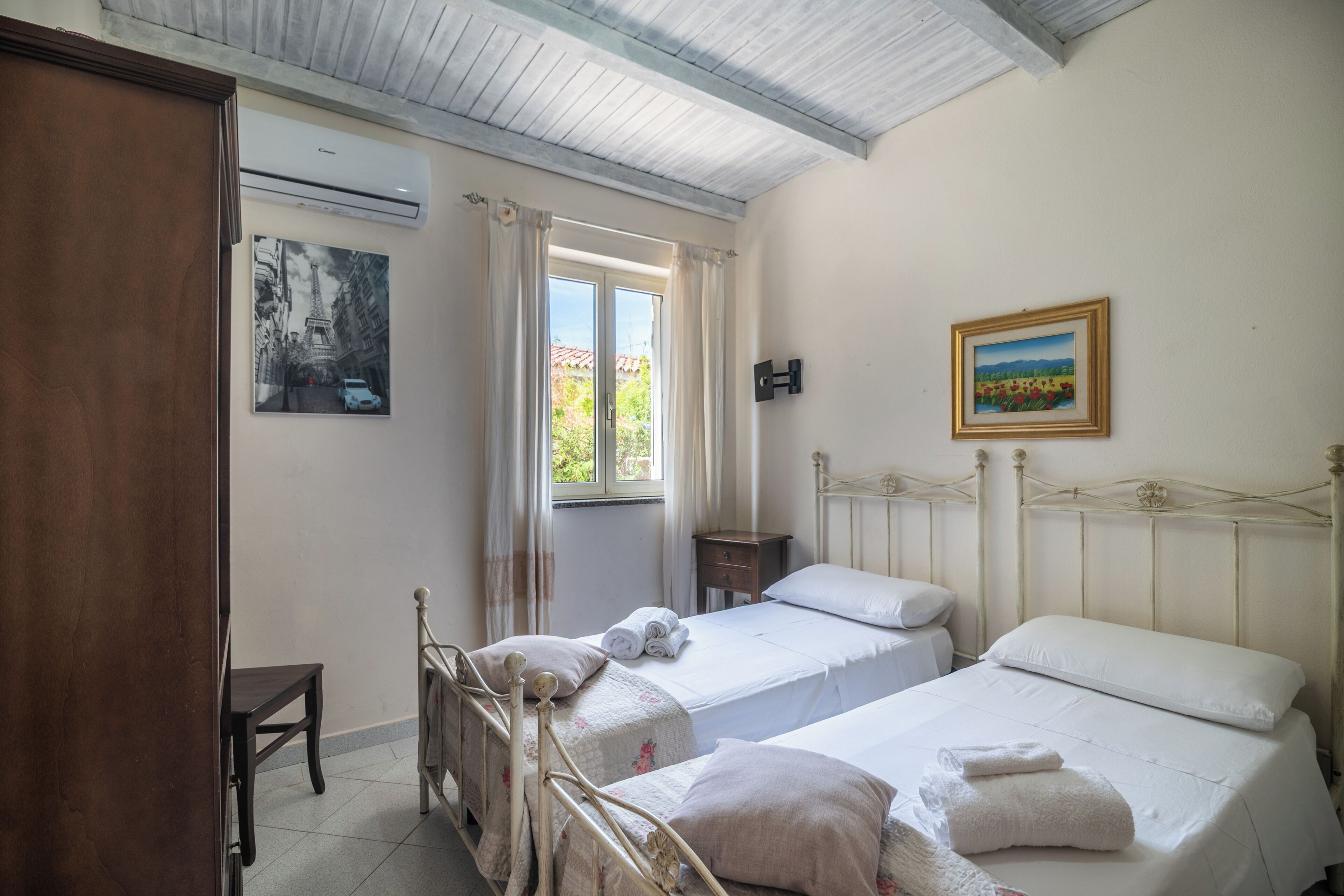 A bedroom with two beds, a wooden wardrobe, a small table, an air conditioning unit, and a painting on the wall.