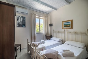 A bedroom with two beds, a wooden wardrobe, a small table, an air conditioning unit, and a painting on the wall. - San Nikola House (Olbia)
