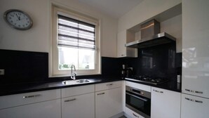A modern kitchen with white cabinets, a black countertop, a stainless steel sink, a window with blinds, a clock, and a stove. - Daelenbroeck Sauna 16 (Susteren)