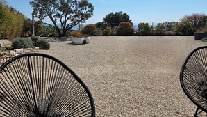 Un área de estar al aire libre con dos sillas de alambre negras, suelo de grava y diversas plantas y árboles al fondo.