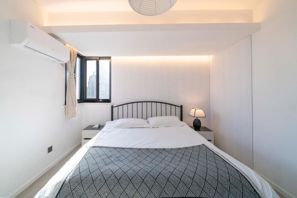 Quarto com cama, mesa de cabeceira, luminária, janela e ar-condicionado.