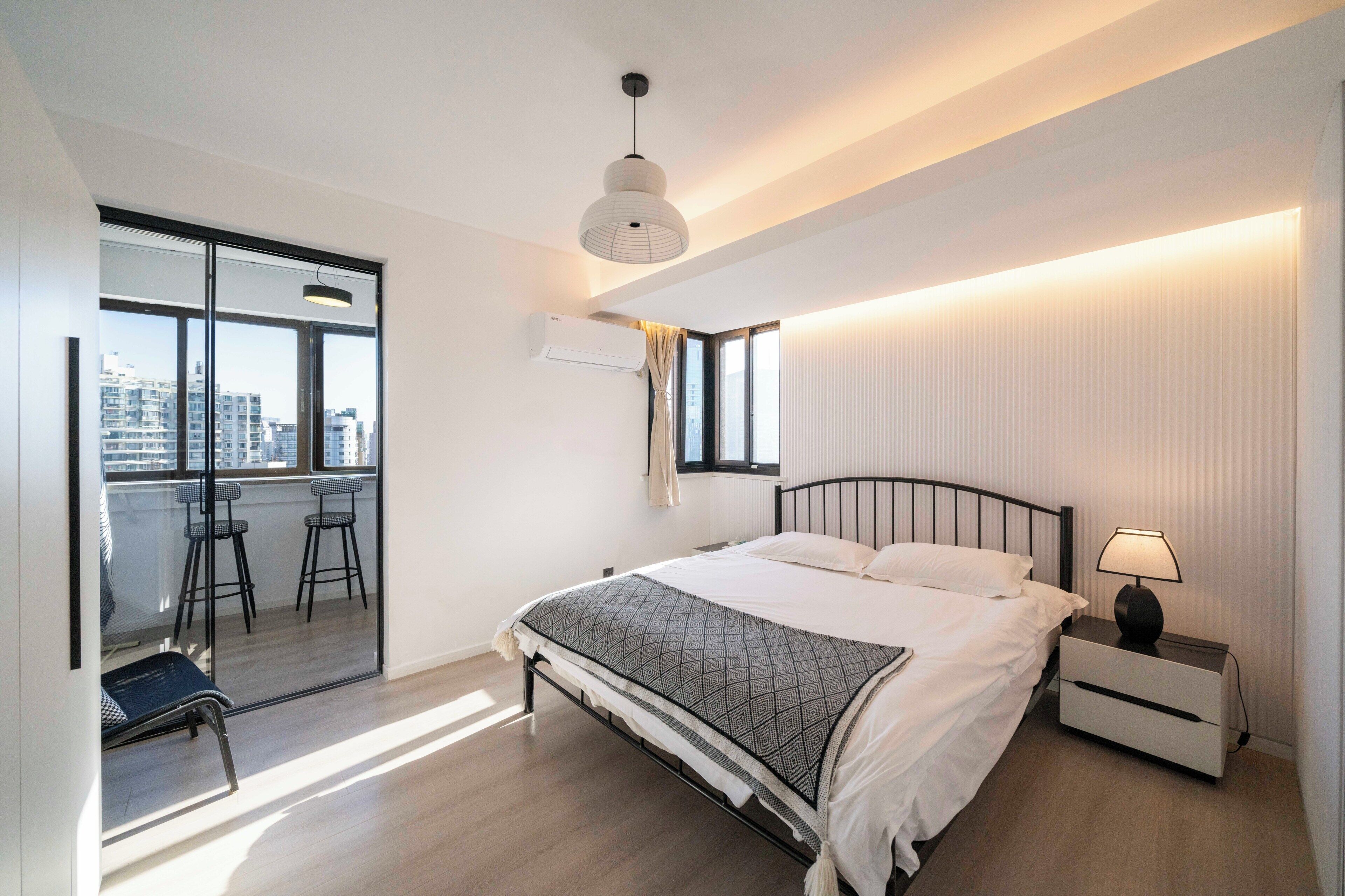 Ein modernes Schlafzimmer mit einem großen Bett, einem Nachttisch, einer Lampe, einem Fenster mit Blick auf Gebäude und einem Balkon mit Sitzgelegenheiten.