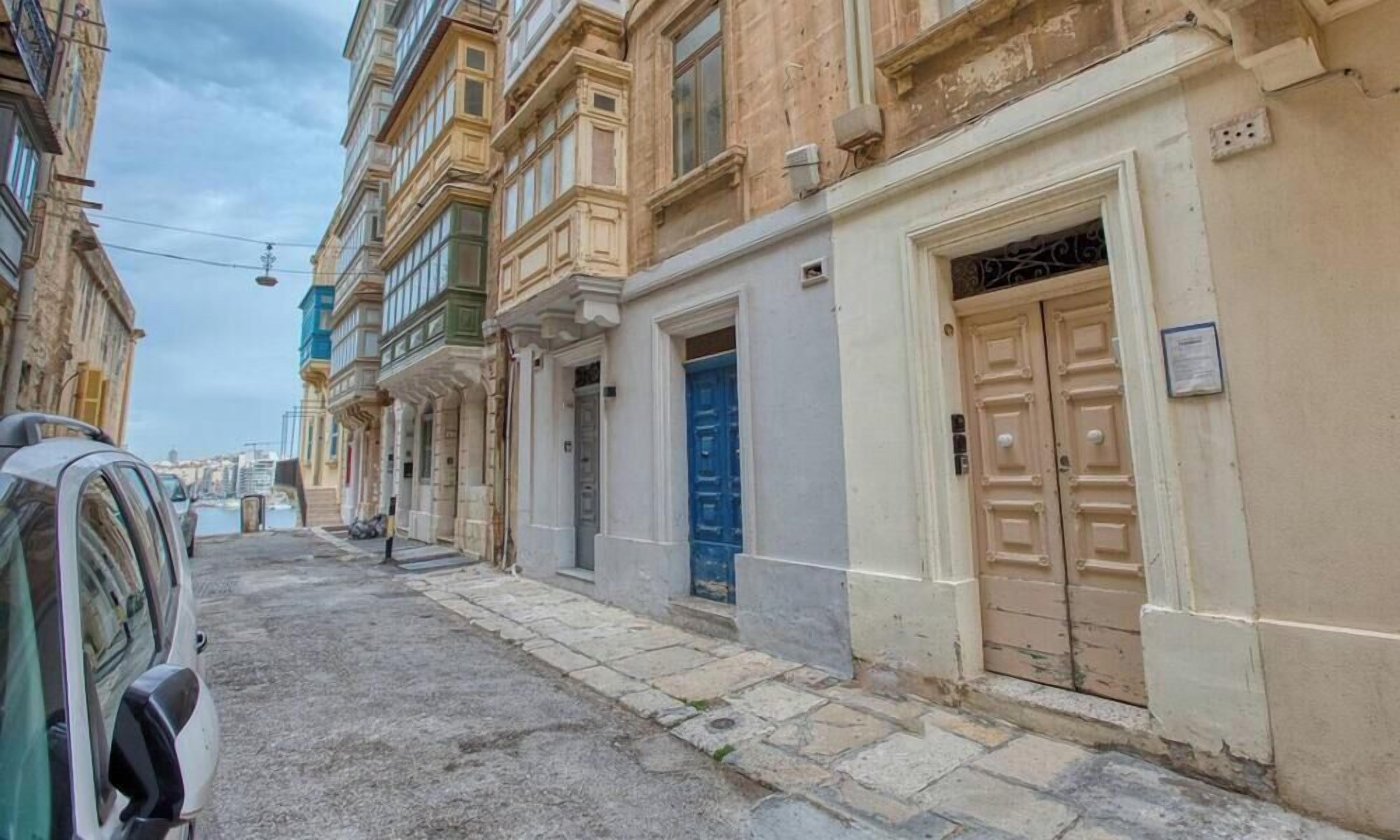 Eine enge Straße mit Häusern an beiden Seiten, die verschiedene Architekturstile zeigen, darunter ein Gebäude mit einer blauen Tür und ein weiteres mit einer beigen Tür.
