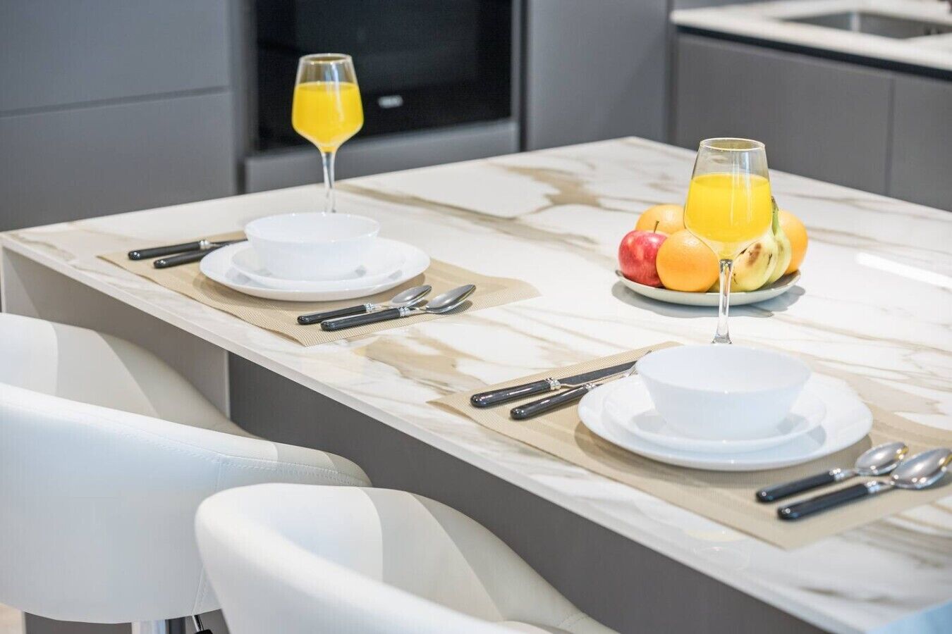 A modern kitchen with a marble countertop, two white plates with cutlery, a glass of orange juice, and a bowl of fruit.