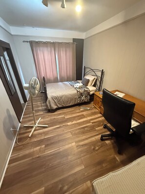 Een slaapkamer met een bed, een bureau, een stoel, een ventilator en een stekkerdoos.