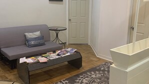 Een woonkamer met een bank, een salontafel met tijdschriften, een kamerkleed met patroon, een witte kast en een deur.