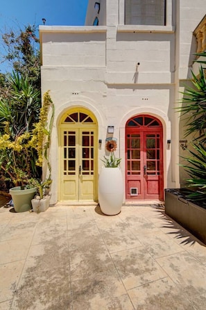 A building with two arched doors, one yellow and one red, and a large white planter in the center. - Gozo Couple Bungalow No 2 · Pool, Cabana, BBQ Deck (Gozo)