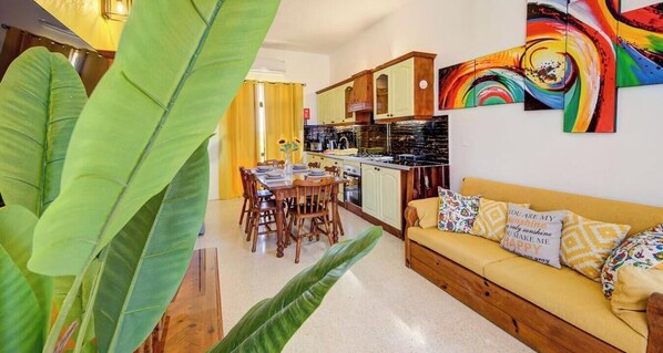 Una cocina con armarios de madera, una zona de comedor con mesa y sillas, y un sofá con cojines estampados.