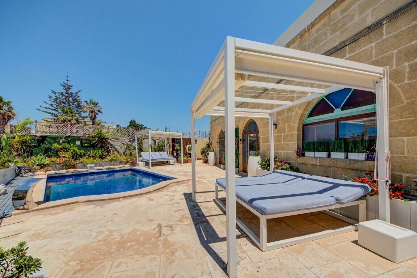 A pool area with a pergola, lounge chairs, and a stone building with arched windows.