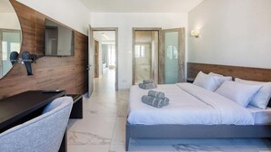 Hotellihuone, jossa on sänky, työpöytä, tuoli, peili ja ovi, joka johtaa toiseen huoneeseen.