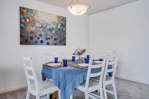Uma área de jantar com mesa redonda, cadeiras brancas, toalha de mesa azul e um quadro em mosaico na parede.