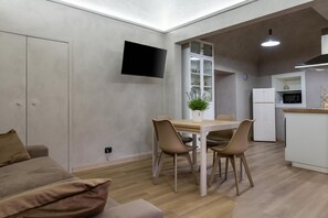 Ein modernes Wohnzimmer mit einer Couch, einem Esstisch mit Stühlen, einem flachbildfernseher an der Wand und einer Küchenzeile inklusive Kühlschrank und Mikrowelle.