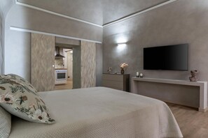 A modern bedroom with a large bed, a flat-screen TV, a dresser, and a kitchen area visible through an open door.
