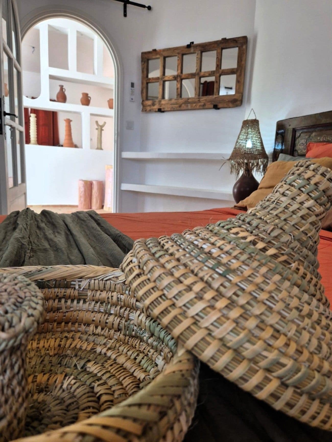 A room with a bed, a woven basket, a shelf with pottery, and a wall-mounted mirror.