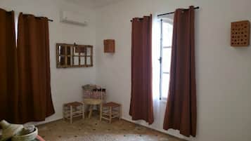 Une pièce avec un plancher en bois, un lit, une fenêtre avec des rideaux marron, un miroir et un petit tabouret.