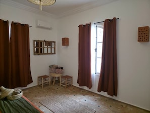 A room with a wooden floor, a bed, a window with brown curtains, a mirror, and a small stool.
