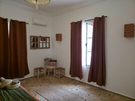 Una habitación con suelo de madera, una cama, un ventanal con cortinas marrones, un espejo y un taburete pequeño.