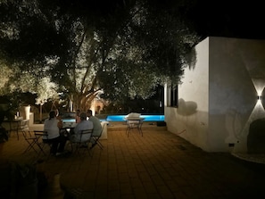An outdoor seating area with tables and chairs, a pool, and a white wall with a light fixture.