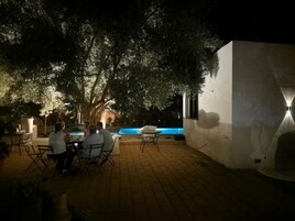 Un espacio exterior con mesas y sillas, piscina y pared blanca con un accesorio de iluminación.