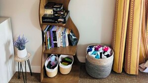 Ein Eckzimmer mit einem hölzernen Bücherregal, einem Bild mit einer Ananas in einem Rahmen, einer kleinen Topfpflanze und einem gewebten Korb.