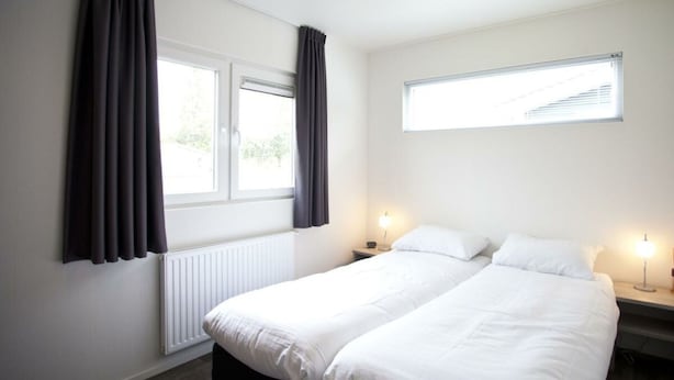 Et hotelværelse med to senge, et vindue med gardiner, en radiator og et lille natbord med lampe.
