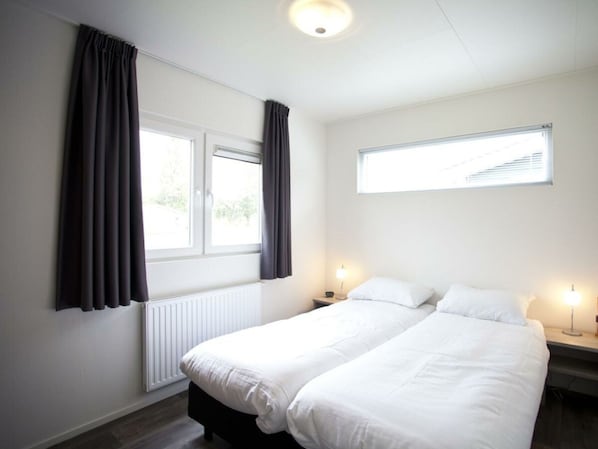A hotel room with two single beds, a window with curtains, a radiator, and a small bedside table with a lamp. - Holiday Home in Nijkerk With Terrace (Nijkerk)