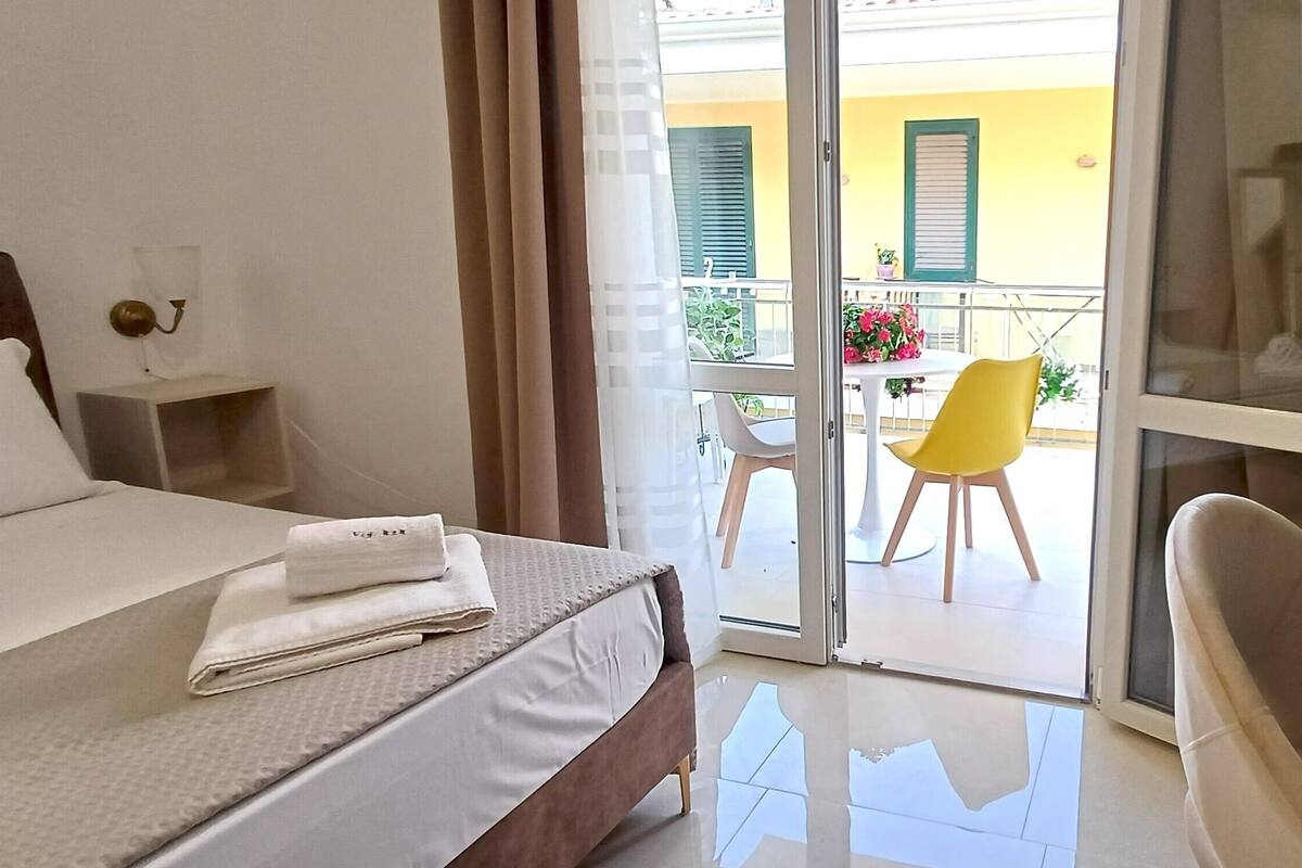 A hotel room with a bed, a desk, a chair, a lamp, and a balcony with a table and chairs.