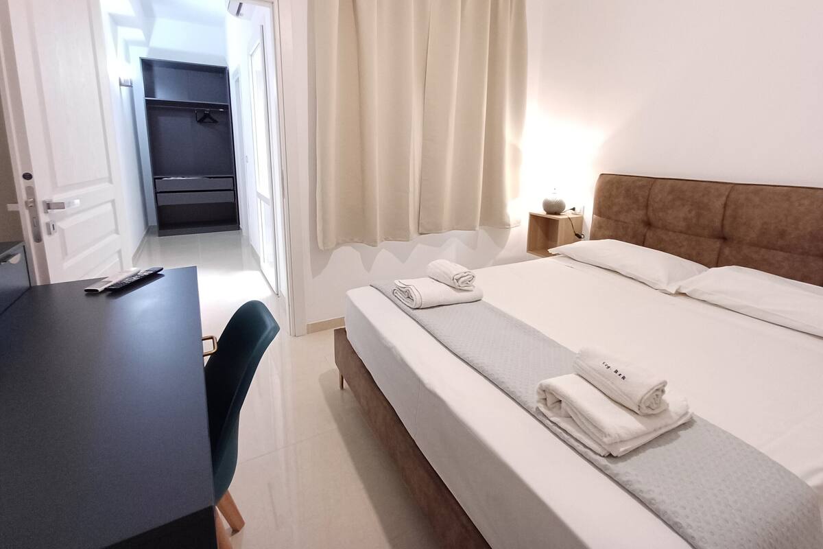 A hotel room with a bed, a desk, a chair, a nightstand with a lamp, and a closet.