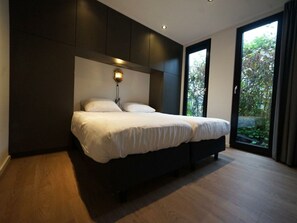 A modern bedroom with a large bed, built-in dark cabinetry, a wall-mounted light, and a large window with a view of greenery. - Holiday Home in Velsen-zuid With Terrace (Velsen-Zuid)
